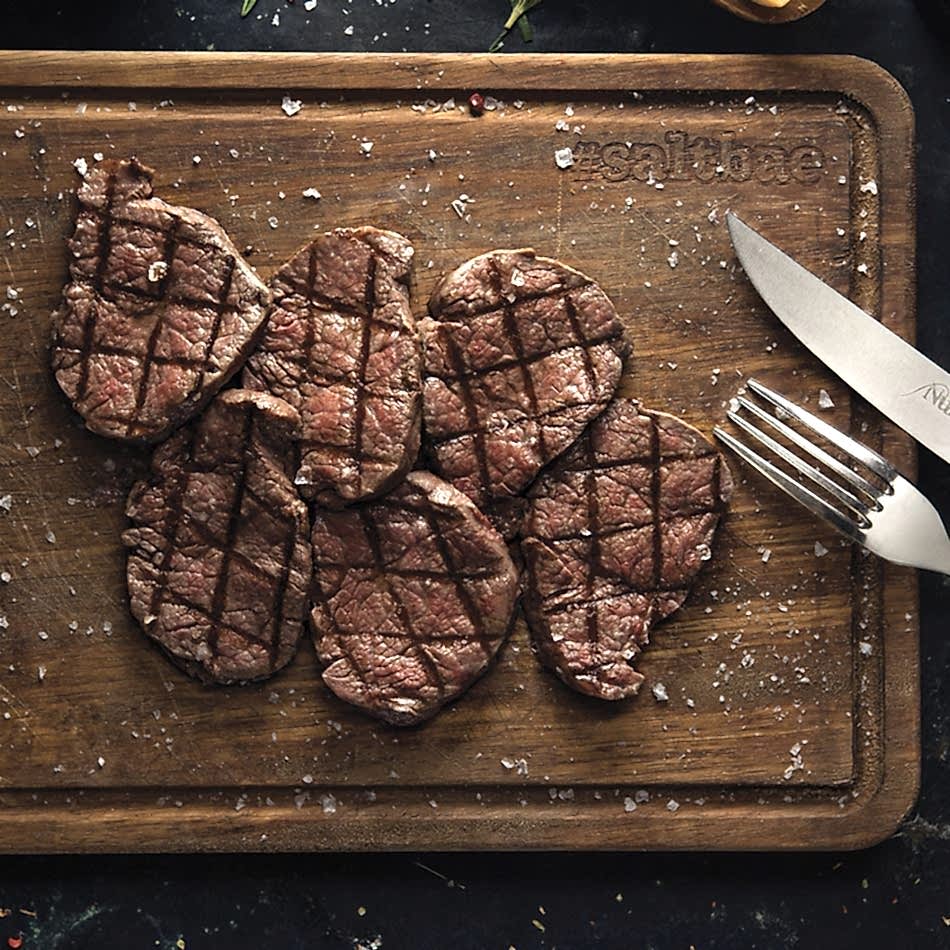 A beautifully presented wooden board featuring grilled wagyu tenderloin slices, arranged neatly with visible grill marks, lightly seasoned with coarse salt, accompanied by a fork and knife engraved with the Nusret logo.
